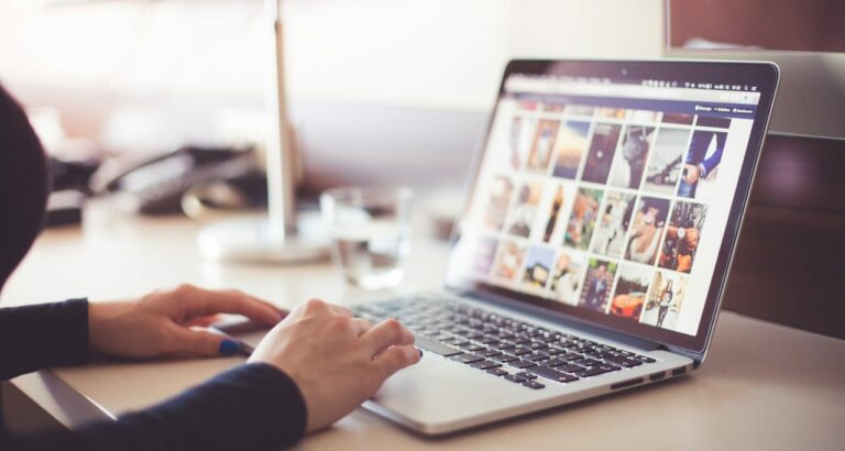 Person using a laptop on a desk with a blurred office environment in the background. The laptop screen displays a grid of various colorful images. The person's hands are on the keyboard and a drinking glass is nearby.