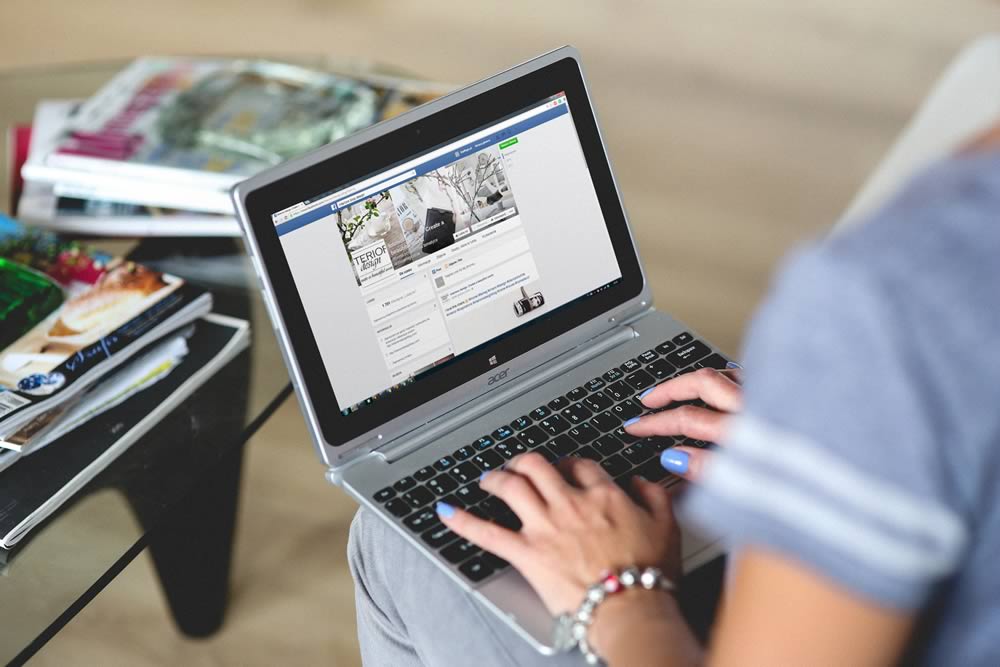 A person using a laptop, browsing a social media profile. Nearby, there are magazines on a glass table. The setting appears to be a casual indoor environment, with the person wearing a gray t-shirt and a bracelet.