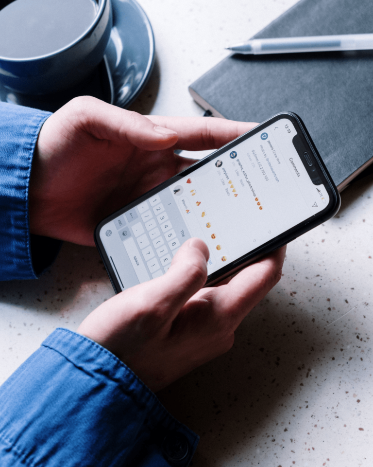 A person wearing a blue jacket is typing on a smartphone, showing a conversation filled with emojis and video media on the screen. A black notebook, a silver pen, and part of a coffee cup are visible on a white speckled surface in the background.