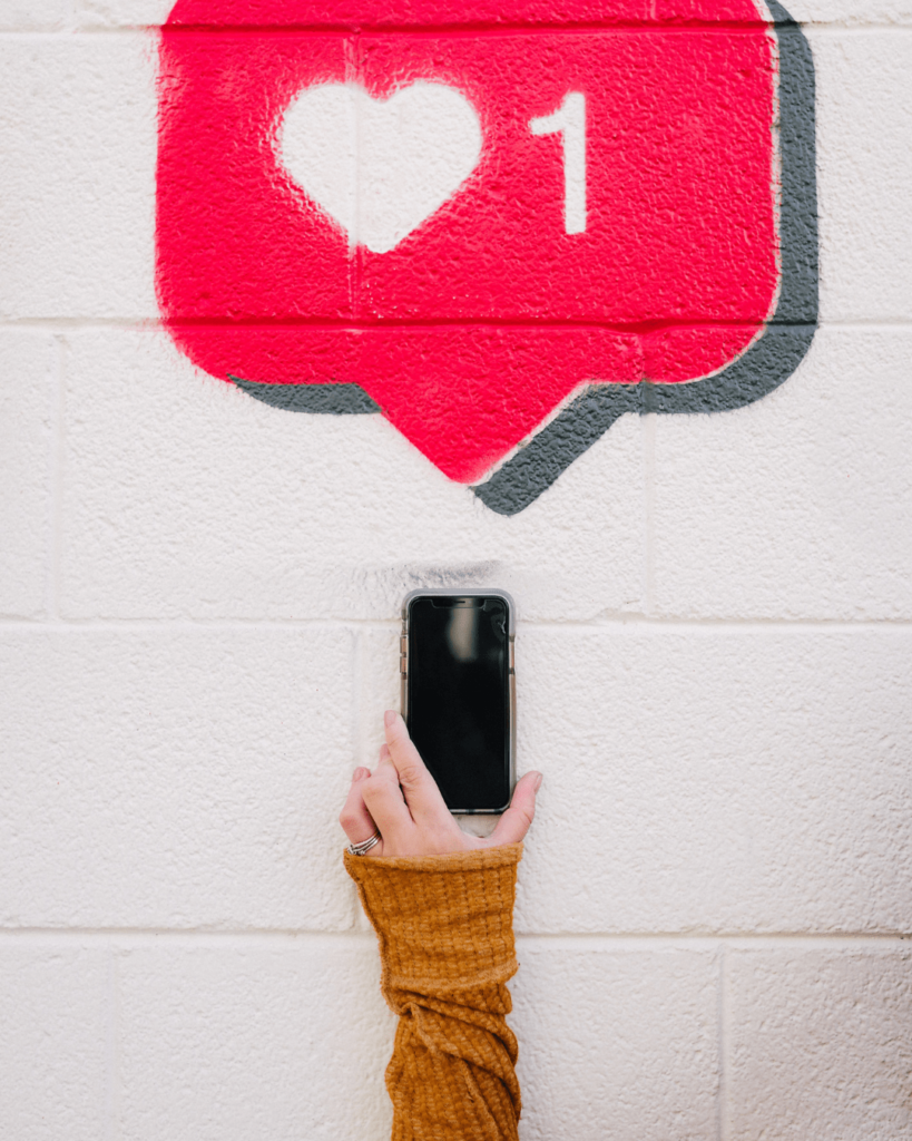 A hand holding a smartphone reaches up towards a painted heart symbol with the number 1 inside, resembling a social media like notification, against a white brick wall.