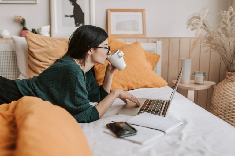 A woman is lying on a bed, propped up on her elbows, using a laptop. She sips from a white mug, surrounded by orange pillows. A notebook and phone are beside her. The room is decorated with framed art and a basket with dried plants.