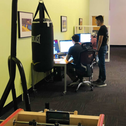 A room with a punching bag and foosball table in the foreground. Three people are seated at desks working on computer screens while one person stands behind them. The walls are light green with framed photos hanging on them.