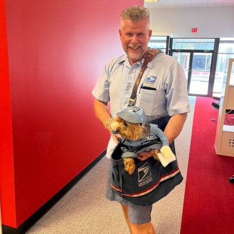 A mail carrier in uniform smiles while holding a small dog dressed in a matching postal outfit, complete with a hat. They are indoors, standing near a red wall and a glass entrance.