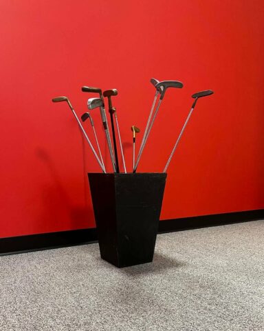 A black container holding multiple golf clubs is placed against a vibrant red wall on a gray carpeted floor. The clubs are arranged with their heads facing outward.