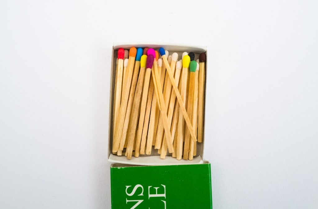 A small box of wooden matches with colorful heads, including red, pink, green, blue, yellow, black, and purple, partially opened on a white surface. The box has a green base with a bit of text visible.
