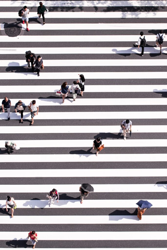 Aerial view of a busy pedestrian crossing with people walking in various directions. The crosswalk consists of wide, alternating black and white stripes. Some individuals carry umbrellas, while others hold bags. Shadows stretch across the striped path.