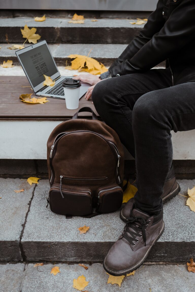 A person dressed in black sits on stairs with a laptop and a disposable coffee cup. A brown backpack is placed on the step next to them. Yellow autumn leaves are scattered on the ground.