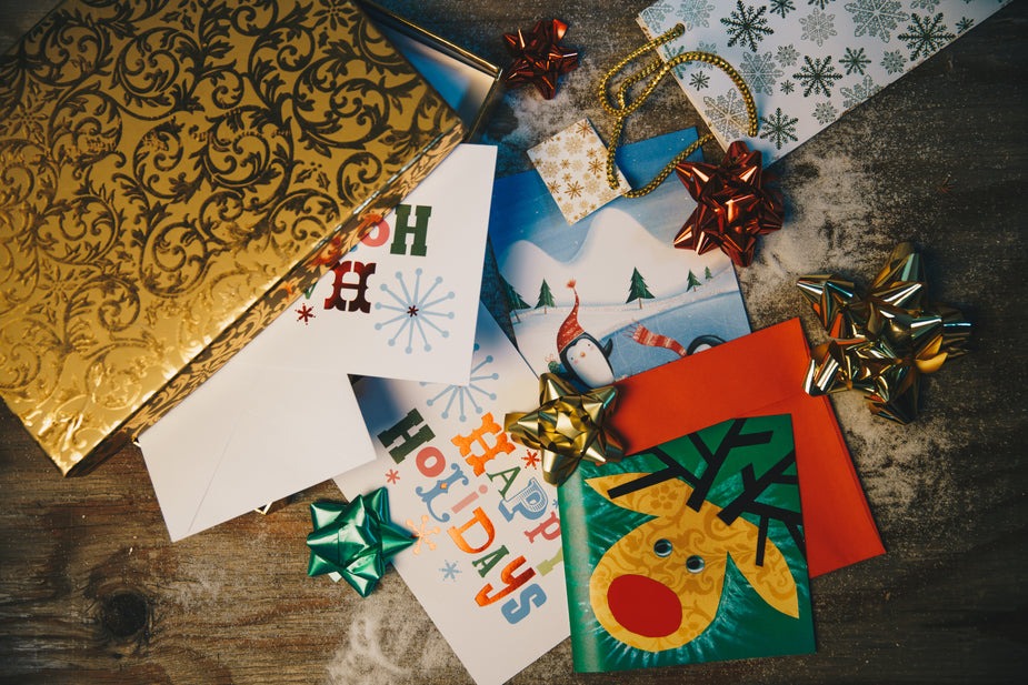 A collection of holiday-themed greeting cards with various designs, including snowflakes, a reindeer, and a snowman. They are surrounded by colorful bows, a golden envelope, and decorative ribbon on a wooden surface dusted with snow.