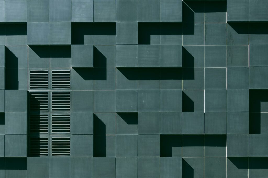 Geometric architectural wall with green square tiles forming a three-dimensional pattern. Rectangular vents are embedded within the tiles, creating a modern and abstract design. Shadows cast varying shapes across the surface.