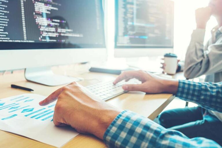 Two people are seated in front of computer monitors filled with lines of code