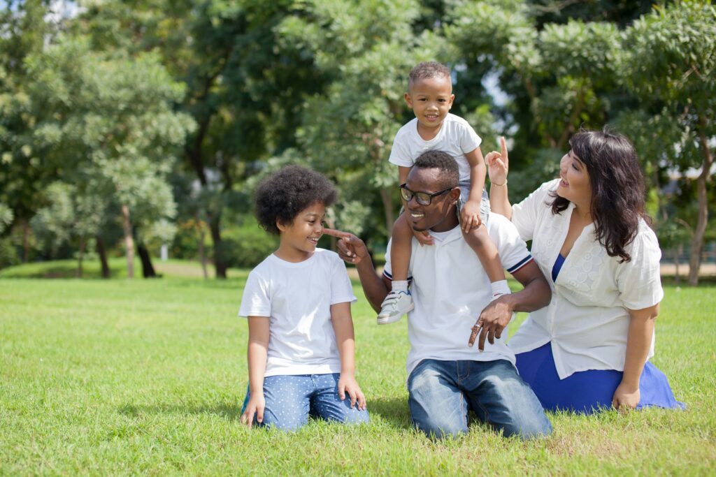 A smiling family of four in a park. A man is kneeling with a toddler on his shoulders, both laughing. A woman sits beside, playfully pointing. A child kneels nearby, grinning. They're surrounded by grass and trees on a sunny day.
