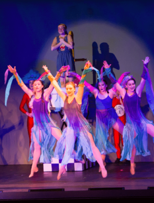 Performers in colorful costumes dance energetically on stage, with one person elevated at the back. The scene is vibrant, featuring purple and blue flowing attire, and the background is lit with a spotlight.