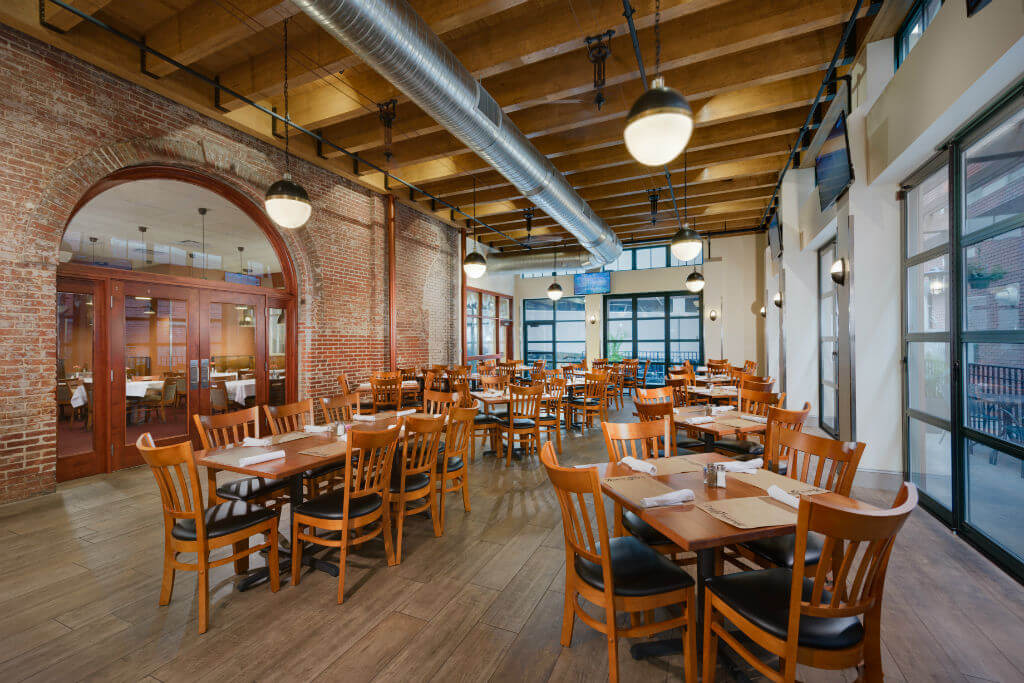 A spacious restaurant interior with wooden tables and chairs. Exposed brick walls and wooden beams create a rustic ambiance. Large windows let in natural light, and pendant lights hang from the ceiling, illuminating the space.