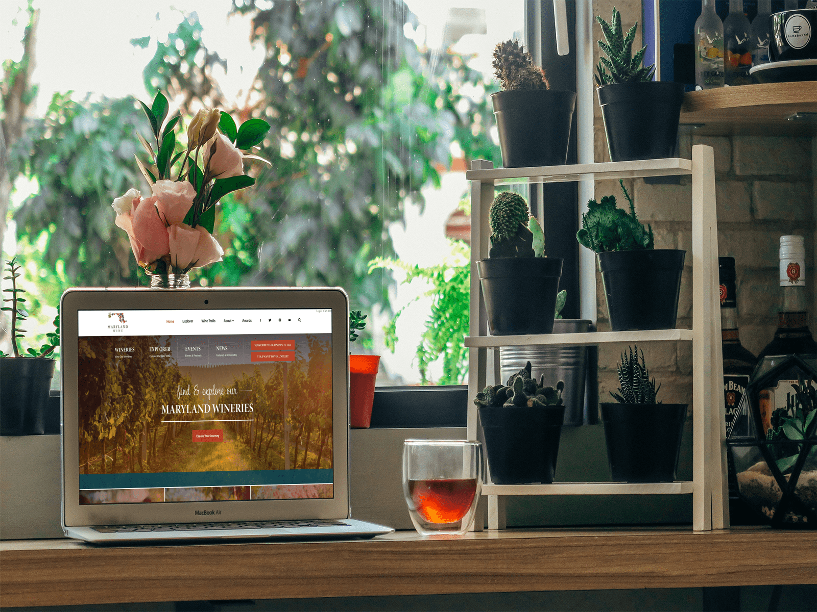 A laptop on a counter displays a vineyard website. Next to it are a glass of tea and a wooden shelf with potted succulents. In the background, a window reveals green foliage and a vase with pink flowers.