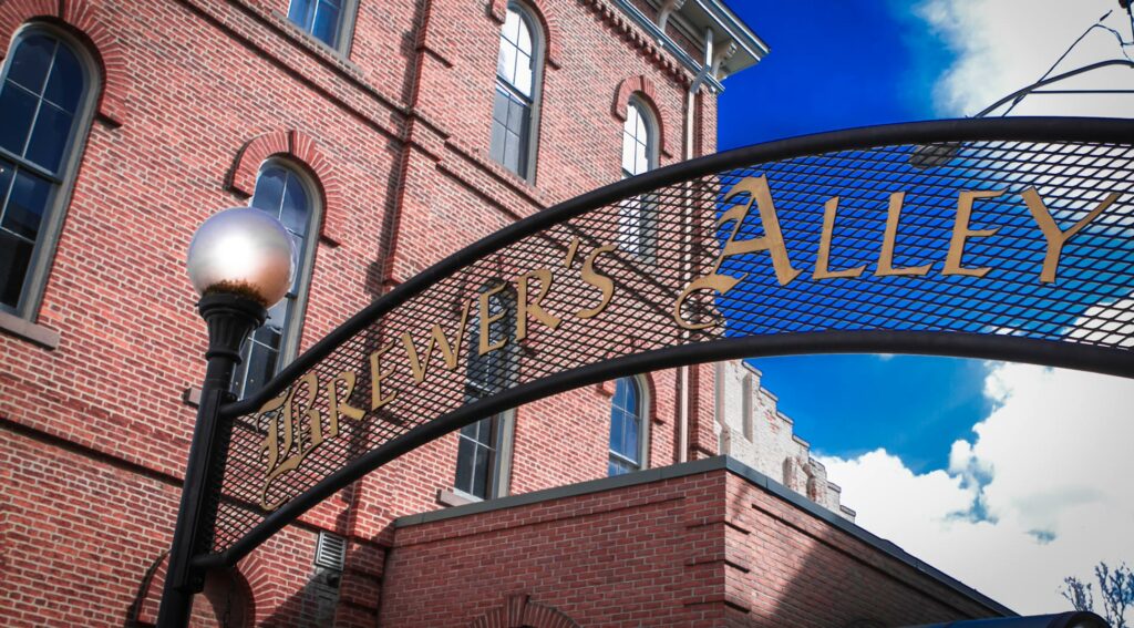 A brick building with arched windows stands under a clear blue sky. In the foreground, a black iron archway displays the words