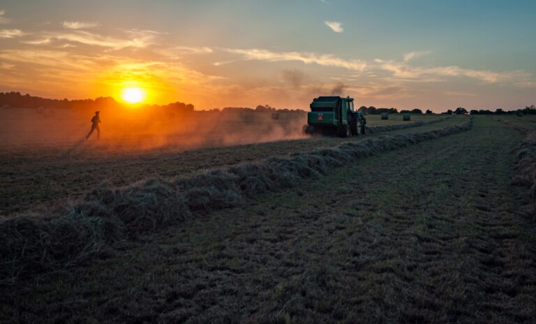 A tractor drives through a grassy field under a sunset sky, creating a trail of dust. A person walks nearby, silhouetted against the bright orange sun. The sky is partly cloudy with shades of blue and orange.