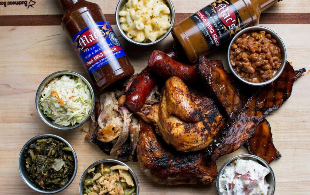 A wooden platter with grilled chicken, sausages, and ribs, surrounded by dishes of coleslaw, baked beans, macaroni and cheese, greens, and potato salad. Two bottles of barbecue sauce are positioned at the top.