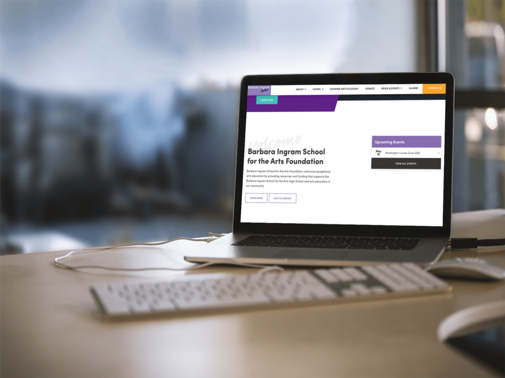 A laptop sits on a desk displaying a website for Barbara Ingram School for the Arts Foundation. The screen shows menu options and a section for upcoming events. A keyboard and mouse are positioned in front of the laptop.