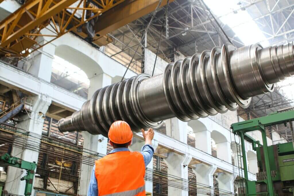 A worker in a safety vest and helmet guides a large, spiral metal shaft being lifted by a crane inside a spacious industrial factory with high ceilings and exposed beams.