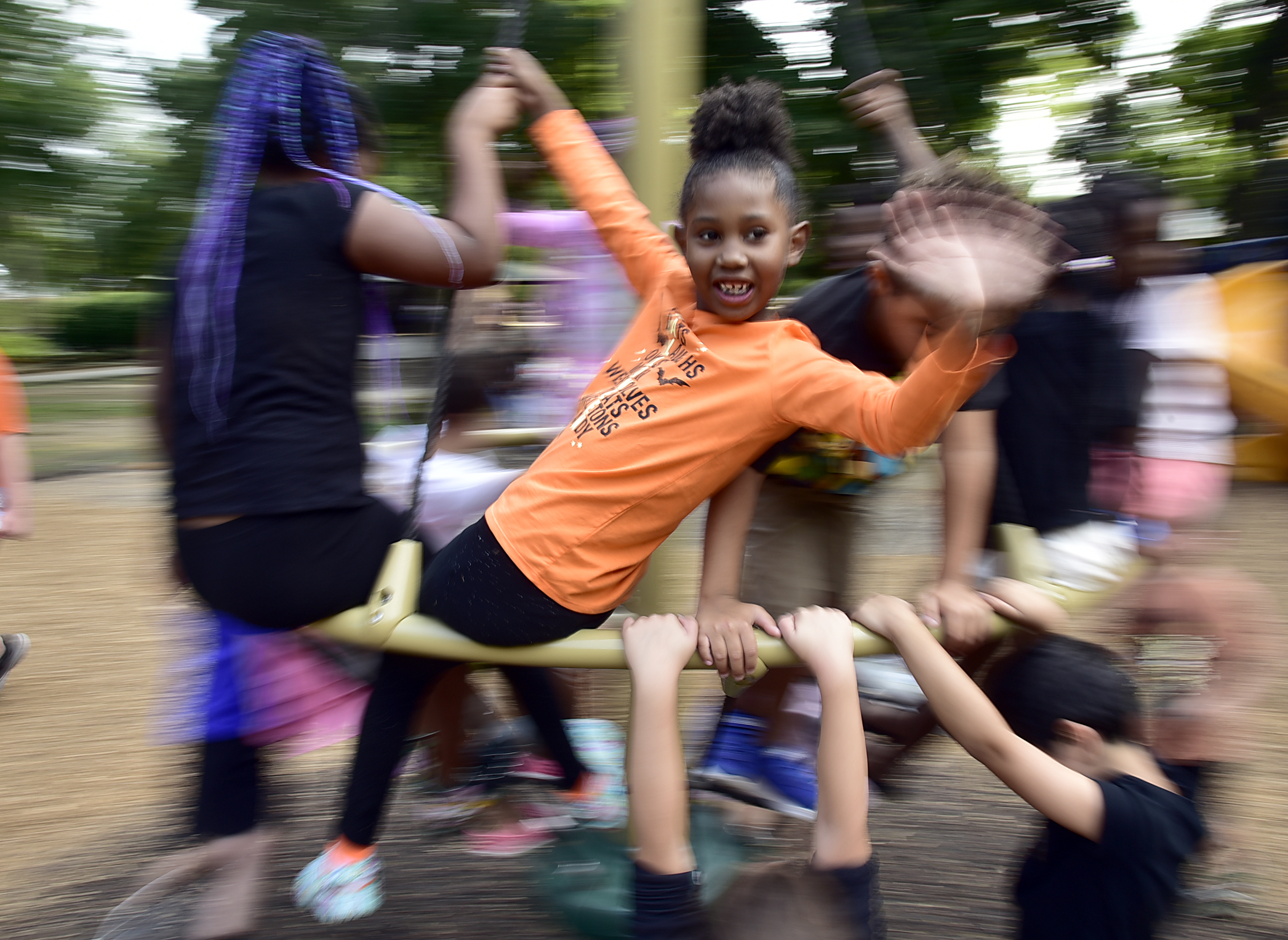 A group of children play energetically on playground equipment. One girl in an orange shirt smiles and waves at the camera while spinning, surrounded by other blurred, moving children.