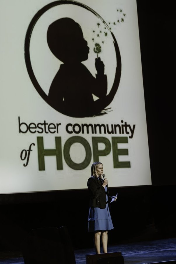 A woman stands on stage speaking into a microphone in front of a large screen displaying the