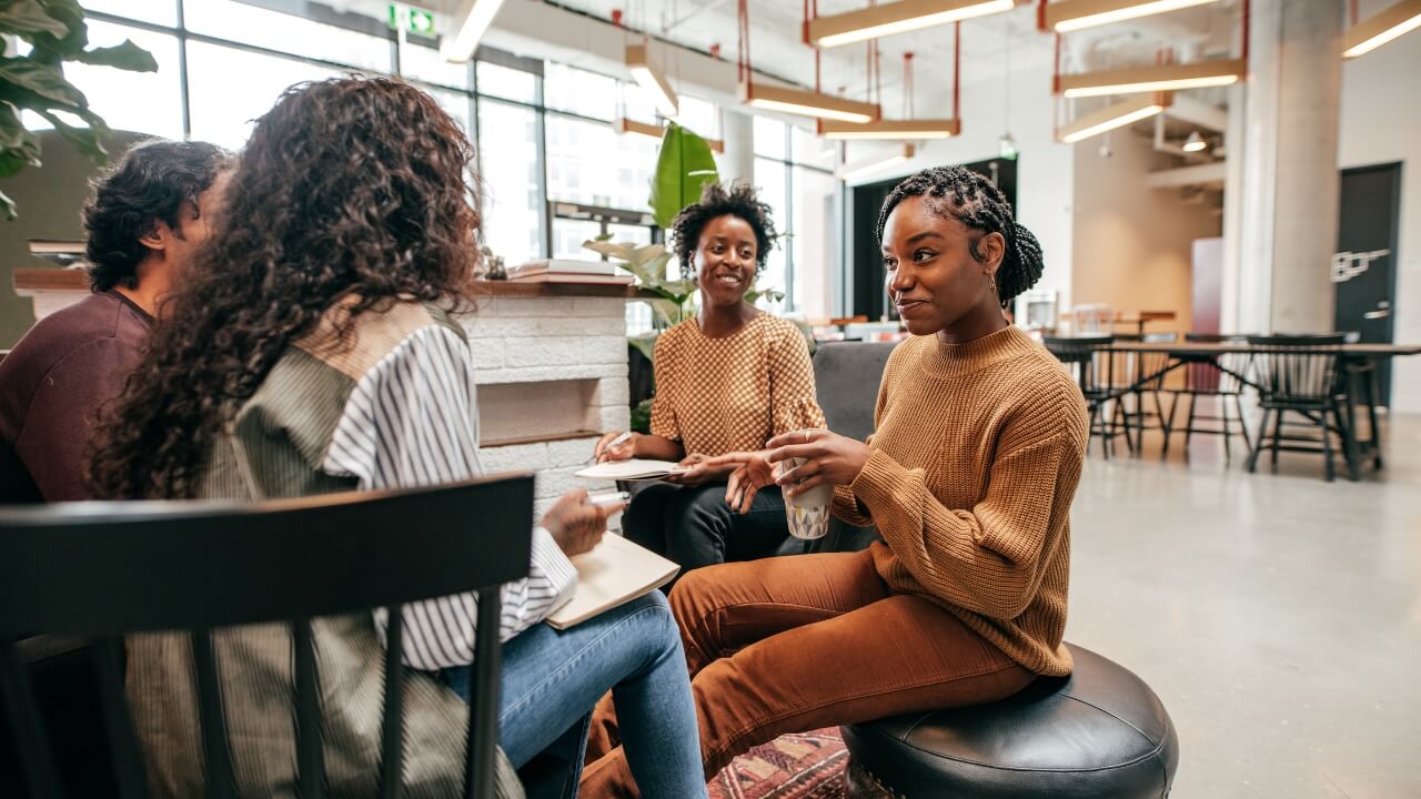 Four people sit together in a modern, well-lit office space, engaged in conversation. Two women face each other, one gesturing while holding a glass, and notebooks are open as they discuss or collaborate.