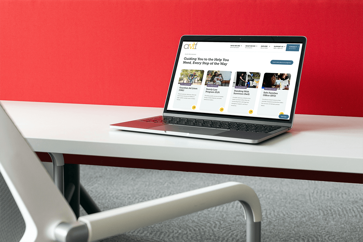 A laptop on a white desk displays a website with articles and images. The desk is next to a white chair, and there is a red wall in the background. The setting appears to be a modern office or workspace.