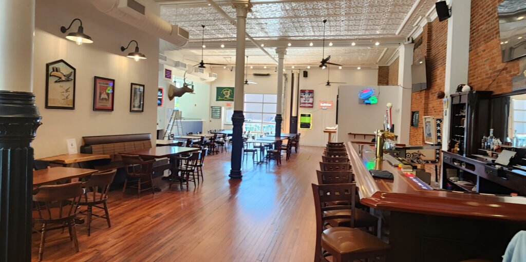 Spacious restaurant interior with wooden floors, tables, and chairs. A bar with stools is on the right, and eclectic wall decorations and neon signs adorn the walls. Large windows at the back let in natural light.