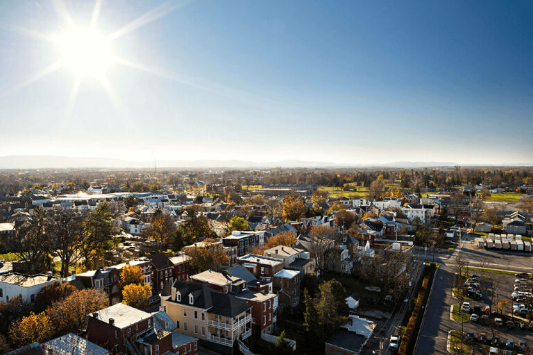 Aerial view of Hagerstown's Bester community, a small neighborhood with houses, trees, and streets under a bright, sunny sky. The sun shines intensely, casting clear shadows, and distant hills are visible on the horizon.
