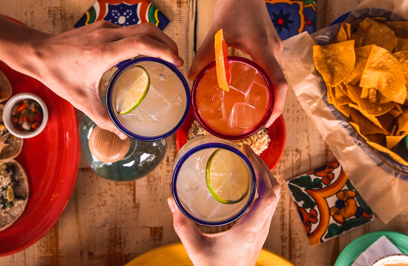 Three hands holding colorful glasses with cocktails garnished with lime and orange, seen from above. Surrounding them are plates of tortilla chips and salsa on a vibrant, festive table.