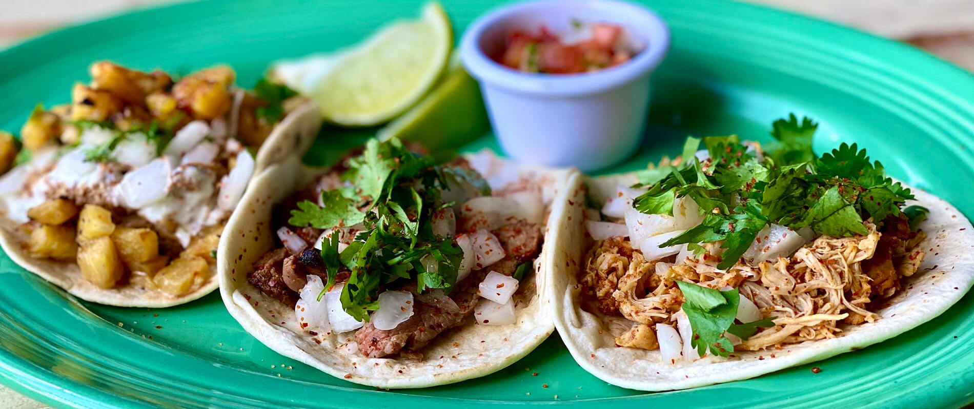 Three tacos filled with various meats, onions, cilantro, and pineapple are arranged on a green plate. A lime wedge and a small cup of salsa are placed in the background.