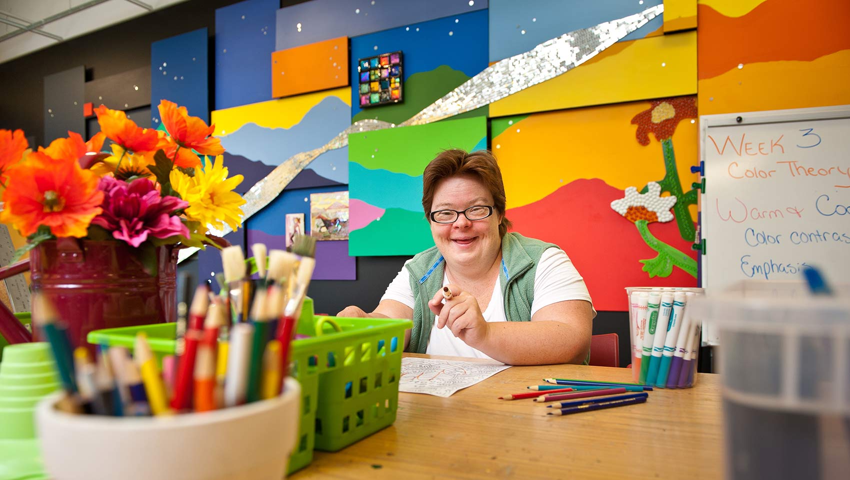 A woman with Down syndrome smiles at a table with art supplies, colorful pencils, and flowers in the foreground. Behind her is a bright, abstract mural and a whiteboard with a lesson on color theory.