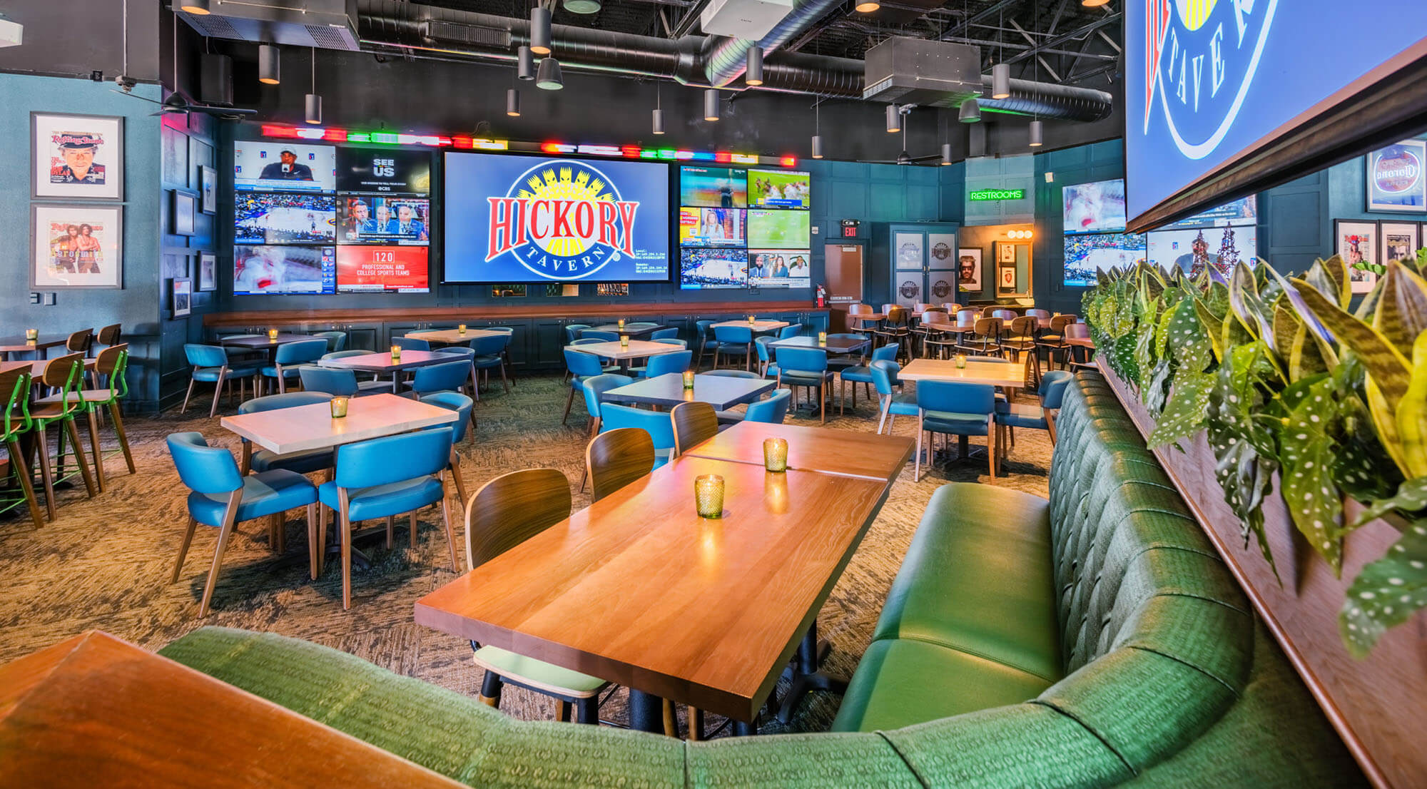 A spacious sports bar with green cushioned booths, wooden tables, blue chairs, potted plants, and large TV screens displaying sports and the