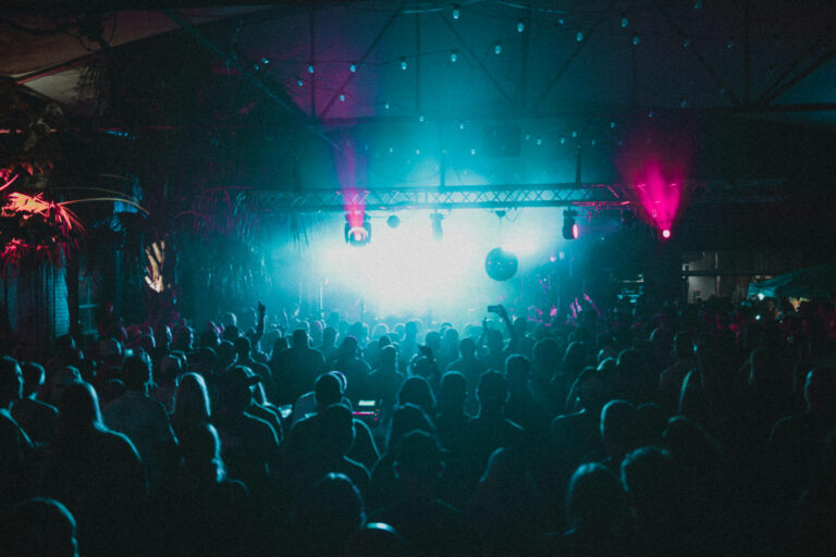 A large crowd is gathered at a concert or music event, facing a brightly lit stage with blue and pink lights, creating a dramatic and energetic atmosphere in a dark indoor venue.