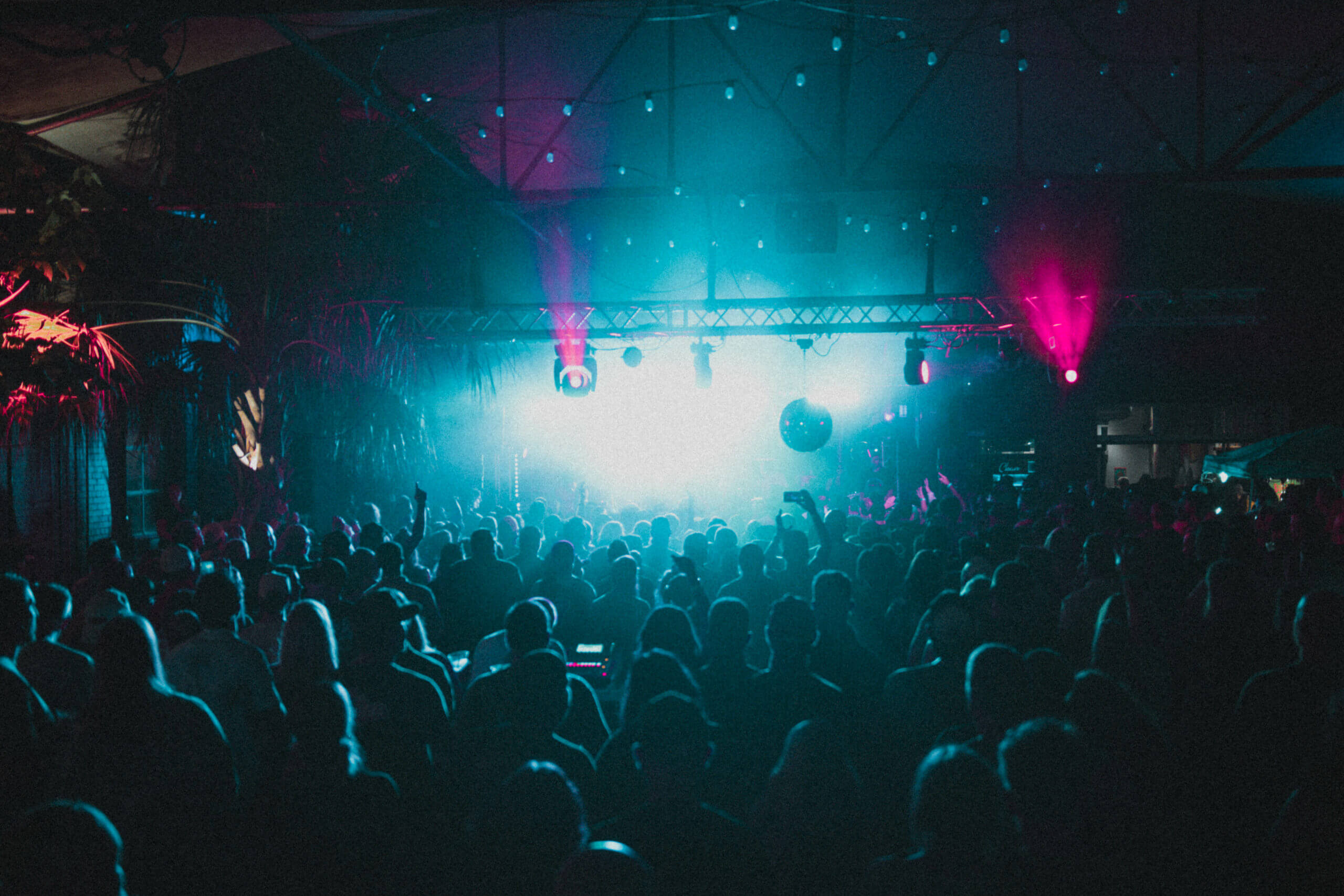 A large crowd is gathered at a concert or music event, facing a brightly lit stage with blue and pink lights, creating a dramatic and energetic atmosphere in a dark indoor venue.