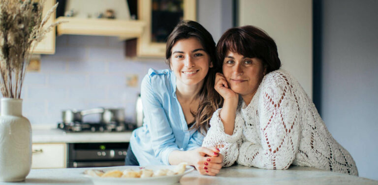 A young woman and an older woman smile as they lean on a kitchen counter together. A vase with dried plants is on the counter, and blurred food is in the foreground. The kitchen setting appears warm and homey.