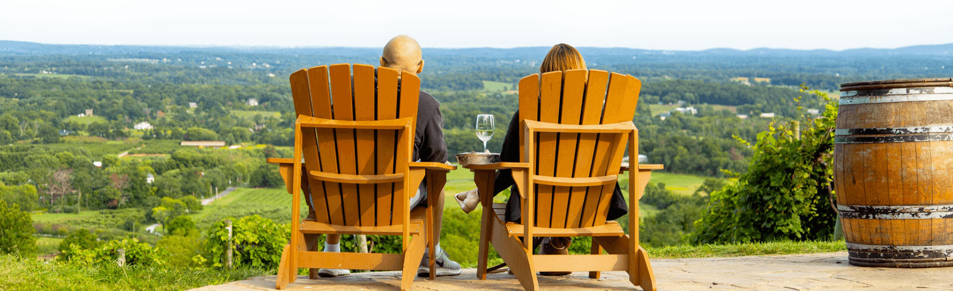 Two people sit in wooden chairs facing a scenic, green valley view with hills in the distance. A wine glass is on a table between them, and a wine barrel stands to the side. The atmosphere is peaceful and relaxing.