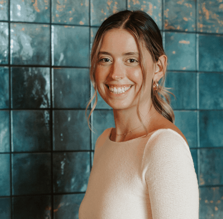 A woman with light brown hair in a low ponytail smiles, wearing a cream-colored long-sleeve top, standing in front of a dark turquoise tiled wall.