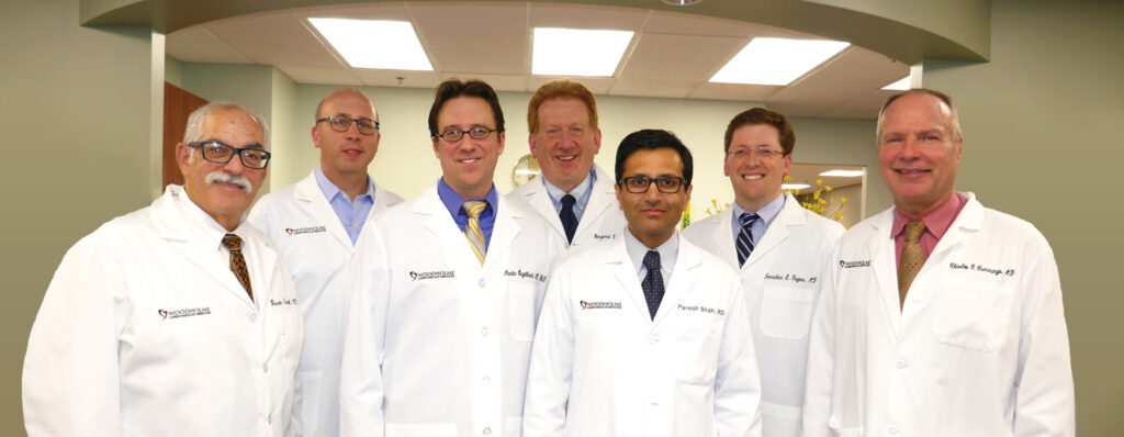 Seven doctors in white lab coats stand together, smiling, in a well-lit medical office with a curved ceiling and fluorescent lights above them.