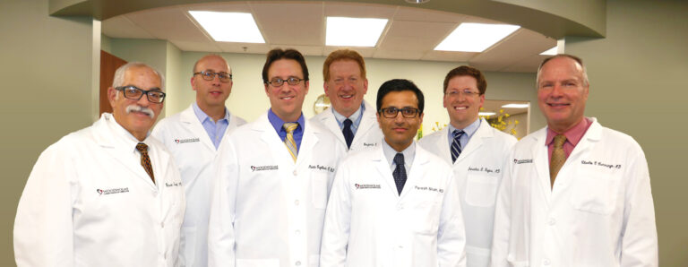 Seven doctors in white lab coats stand together, smiling, in a well-lit medical office with a curved ceiling and fluorescent lights above them.