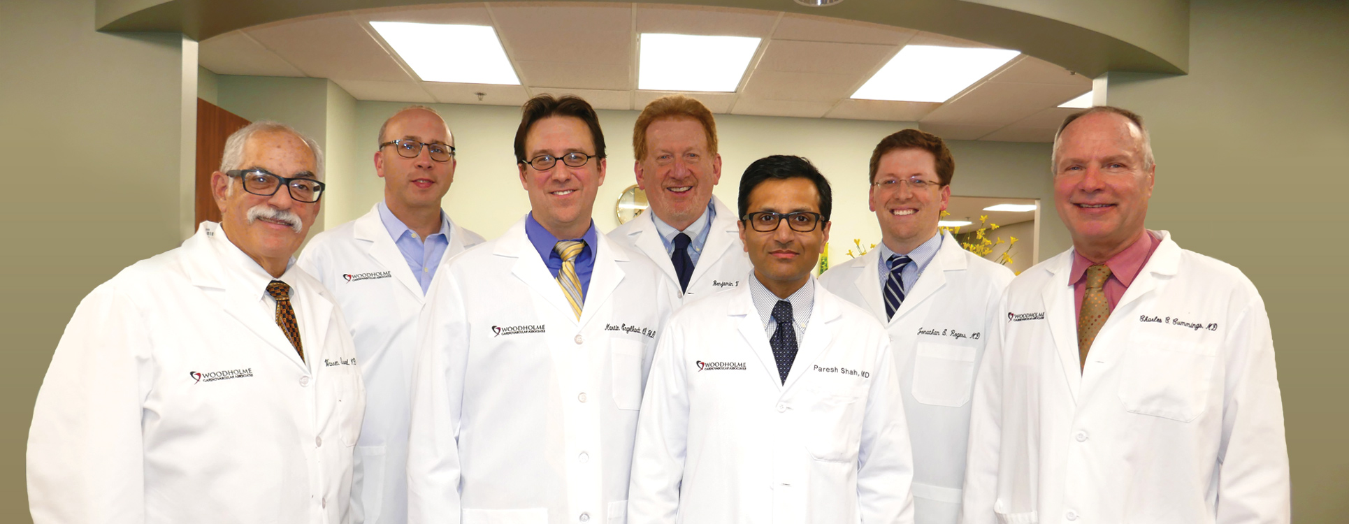 Seven doctors in white lab coats stand together, smiling, in a well-lit medical office with a curved ceiling and fluorescent lights above them.