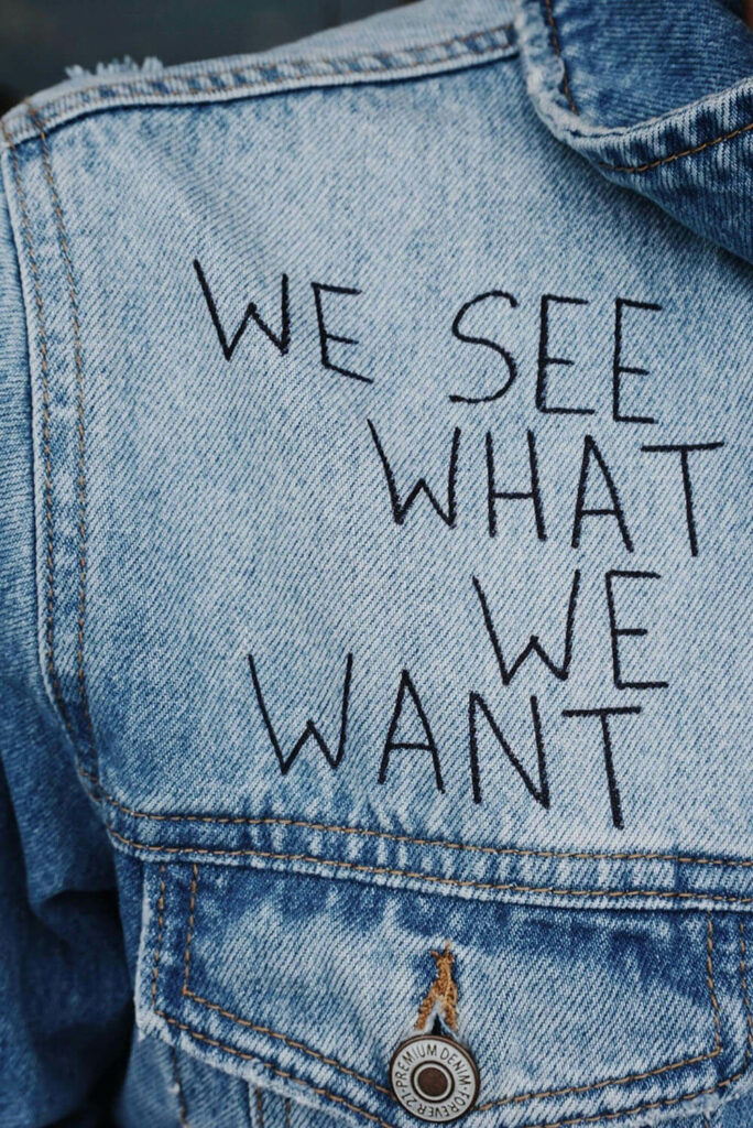 A close-up of the back of a blue denim jacket with the words "WE SEE WHAT WE WANT" stitched in black thread above a buttoned pocket.
