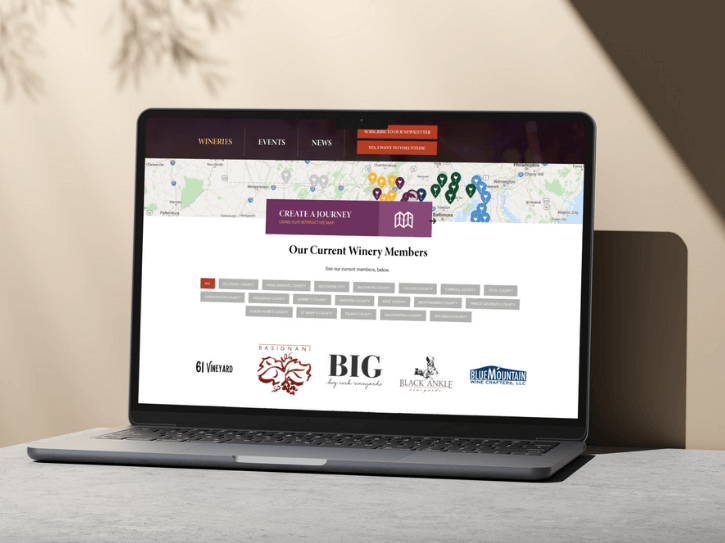 A laptop on a beige surface displays a winery website showing a map, navigation menu, and a list of winery members with logos and gray buttons. Sunlight casts shadows on the wall in the background.