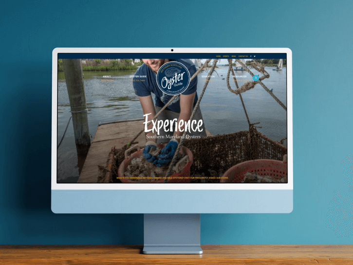 A computer monitor on a wooden desk displays a website featuring Southern Maryland Oysters, with a background image of oysters being harvested on the water. The words “Experience Southern Maryland Oysters” are visible on the screen.