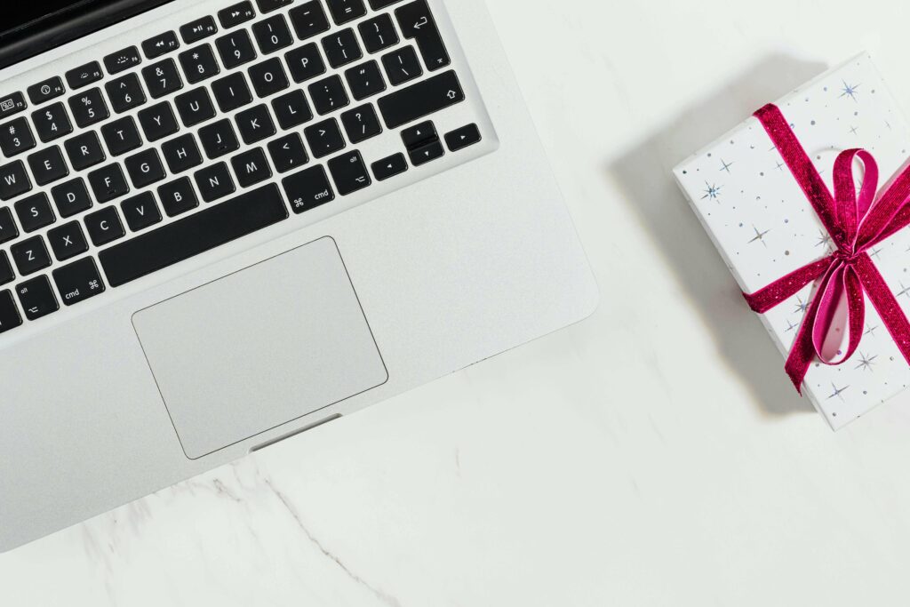 A silver laptop sits on a white marble surface next to a small, white gift box wrapped with a red ribbon and decorated with star patterns.