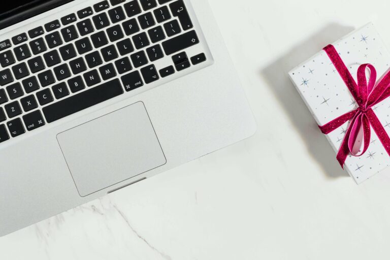 A silver laptop sits on a white marble surface next to a small, white gift box wrapped with a red ribbon and decorated with star patterns.